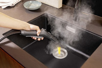 Hand using Kärcher steam cleaner on kitchen sink drain, emitting steam.