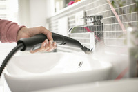Hand using Kärcher steam cleaner on bathroom tap, with steam visible, in a tiled indoor setting.