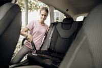Person cleaning car seat with Kärcher vacuum, wearing a striped shirt, in an outdoor setting with trees visible through the window.