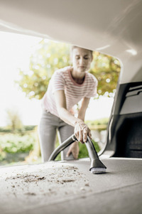 Person using a vacuum cleaner to remove dirt from a car boot, with a blurred background of greenery outside.
