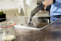 Hand using Kärcher steam cleaner on a stovetop, emitting steam. Indoor kitchen setting with jar and blurred background.