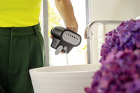 Person in yellow top and green trousers using a Kärcher steam cleaner over a white basin, with purple flowers nearby.