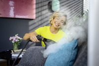 Person using a Kärcher steam cleaner on a grey sofa, with a turquoise cushion. Indoor setting with a pink artwork and flowers in the background.