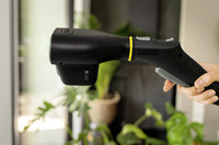 Hand holding a Kärcher steam cleaner nozzle indoors, with blurred plants in the background.