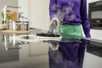 Person using a Kärcher steam cleaner on a kitchen hob, with steam visible. Stainless steel pot and flowers in the background.