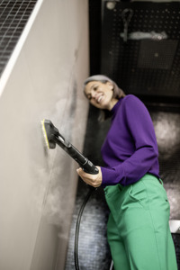 Person using a Kärcher steam cleaner on a tiled wall, wearing a purple jumper and green trousers in an indoor setting.