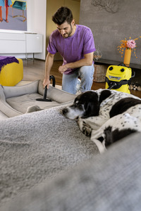 Man using Kärcher vacuum cleaner on dog bed indoors, with a sleeping black and white dog nearby. Bright room with modern decor.
