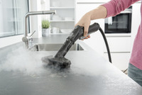 Hand using Kärcher steam cleaner on kitchen countertop, emitting steam near sink in modern kitchen setting.