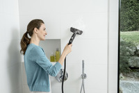 Person using a Kärcher steam cleaner on a white tiled bathroom wall, with toiletries on a shelf and a showerhead visible.