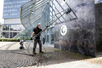 Person cleaning building facade with Kärcher high-pressure cleaner, standing on wet cobblestone pavement outside a modern glass structure.