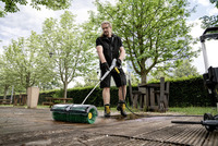 Person using a Kärcher high-pressure cleaner with a brush attachment on a wooden deck in a garden setting with trees and outdoor furniture.