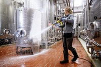 Person using Kärcher high-pressure cleaner to wash large stainless steel tanks in an industrial setting with red tiled floor.