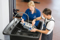 Two people cleaning an engine part in a workshop using brushes and a water basin.