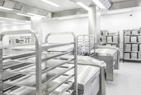 Industrial kitchen with stainless steel racks and trays, large appliances, and stacks of trays on shelves in a bright, clean setting.