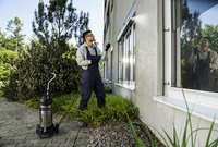 Person cleaning windows with Kärcher high-pressure cleaner outdoors, surrounded by greenery and a building.
