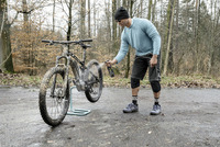 Person cleaning a muddy mountain bike with a spray bottle in a forest setting, wearing a blue top and black shorts.