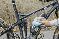 Hand cleaning a muddy black mountain bike frame with a blue cloth outdoors.