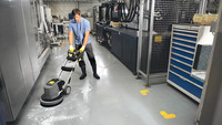 Person using a Kärcher floor scrubber in an industrial setting, surrounded by machinery and storage units.