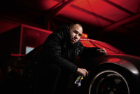 Person in dark clothing crouched beside a car with a red-lit background, holding a bottle of tyre gloss near the wheel.