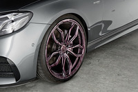 Close-up of a grey car's front wheel with purple cleaning solution on the alloy rim, parked on a concrete surface.