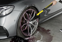Hand cleaning a car wheel with a Kärcher brush, soap and water on the ground, grey car parked indoors.