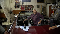 Man in plaid shirt sits in a red convertible inside a garage, surrounded by tools and equipment. A Kärcher product and cloth rest on the car.
