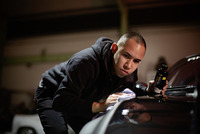 Person polishing a car bonnet indoors, holding a bottle of car care product in one hand and a cloth in the other.
