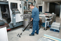 Person using Kärcher industrial vacuum cleaner in a factory setting, cleaning debris from the floor near machinery and metal racks.