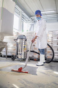 Person in lab coat and boots using a Kärcher industrial vacuum cleaner in a warehouse with stacked bags and machinery.